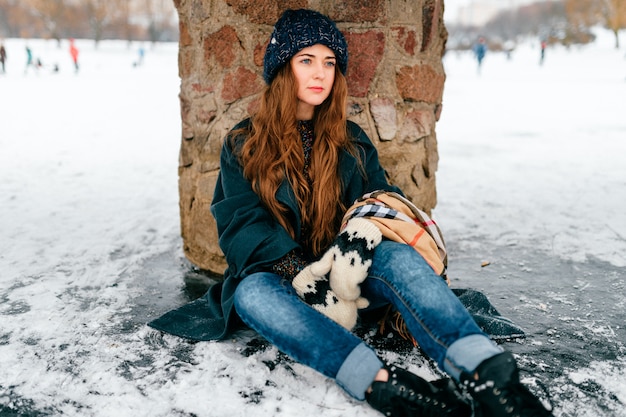 Giovane bella donna in vestiti alla moda con capelli marroni lunghi che si siedono sotto il ponte sul ghiaccio sul lago congelato nel giorno di inverno freddo del gelo