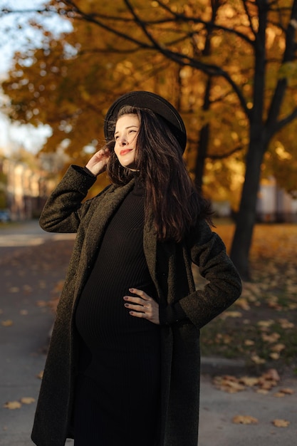 Giovane bella donna incinta con un cappello con i capelli scuri in un vestito e un cappotto attillati neri