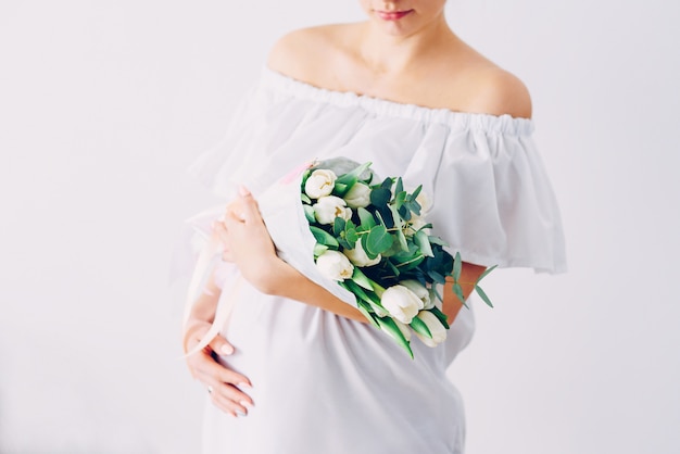 Giovane bella donna incinta in un abito bianco con un mazzo di tulipani bianchi