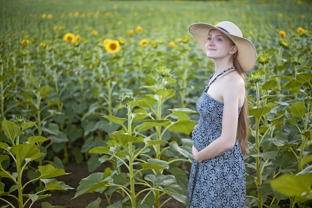 Giovane bella donna incinta si trova in un cappello e un vestito sul campo di girasoli in fiore