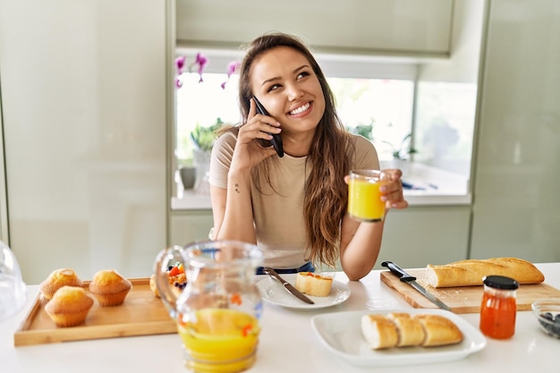 Giovane bella donna ispanica che fa colazione parlando sullo smartphone in cucina