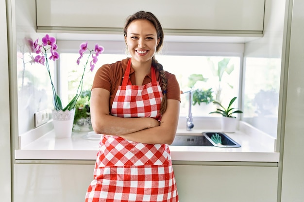 Giovane bella donna ispanica che indossa un grembiule in piedi con le braccia incrociate gesto in cucina