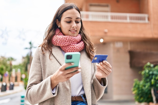 Giovane bella donna ispanica che utilizza smartphone e carta di credito che indossa sciarpa in strada