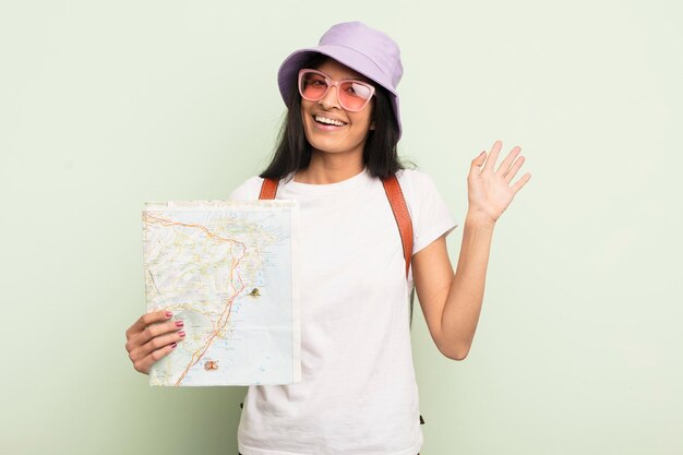 Giovane bella donna ispanica sorridente felicemente agitando la mano dandoti il benvenuto e salutandoti turista e concetto di mappa