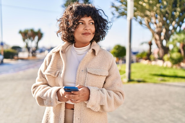 Giovane bella donna ispanica sorridente fiducioso utilizzando lo smartphone al parco