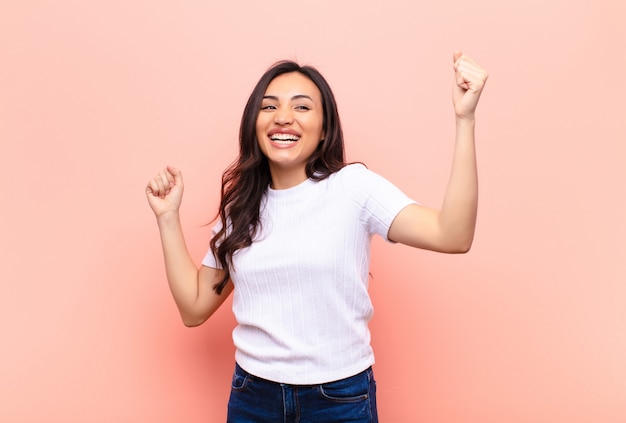 Giovane bella donna latina che grida trionfalmente, guardando come eccitato, felice e sorpreso vincitore, festeggia contro la parete piatta