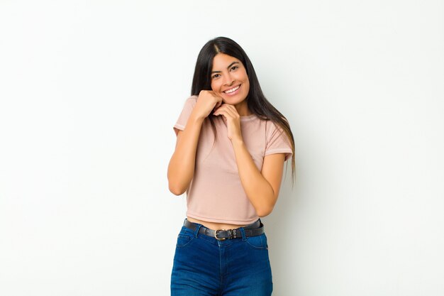 Giovane bella donna latina sentirsi innamorati e sembrare carina, adorabile e felice, sorridendo romanticamente con le mani accanto al viso contro la parete piatta