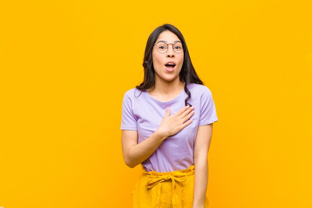 Giovane bella donna latina sentirsi scioccata e sorpresa, sorridendo, prendendosi per mano, felice di essere quella o mostrando gratitudine contro il muro arancione