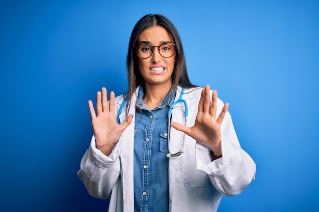 Giovane bella donna medico che indossa stetoscopio e occhiali su sfondo blu impaurito e terrorizzato con il gesto di arresto dell'espressione di paura con le mani che gridano in stato di shock Concetto di panico