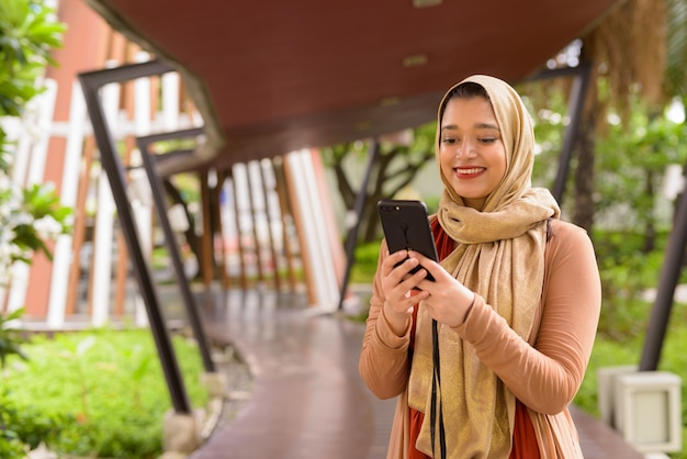 Giovane bella donna musulmana indiana felice che utilizza telefono nella città con la natura