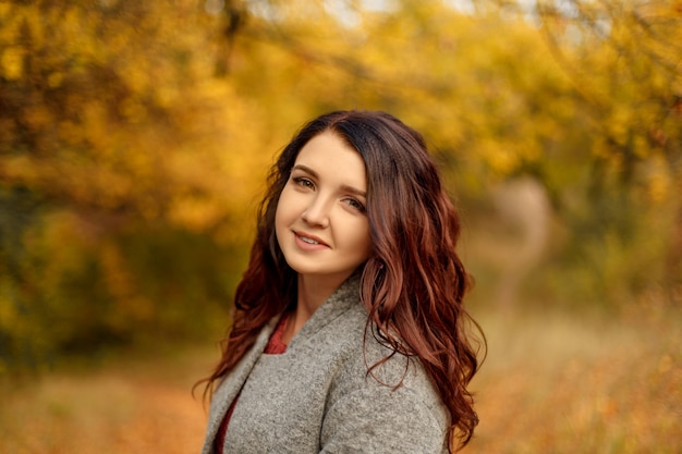 Giovane bella donna nello sweather grigio del cappotto che cammina nel parco di autunno con le foglie gialle e rosse