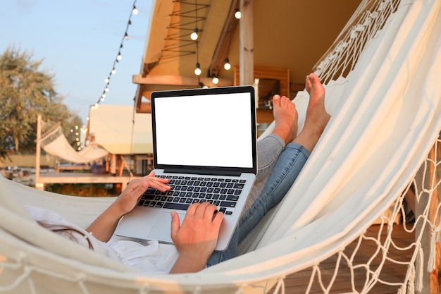 Giovane bella donna sdraiata su un'amaca con un computer portatile al resort. Mock up, schermo bianco vuoto.