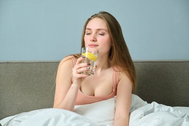 Giovane bella donna seduta a casa a letto con un bicchiere d'acqua al limone