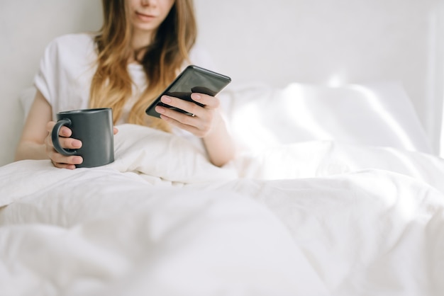 Giovane bella donna seduta a letto e navigare in Internet in uno smartphone con una tazza di caffè in mano