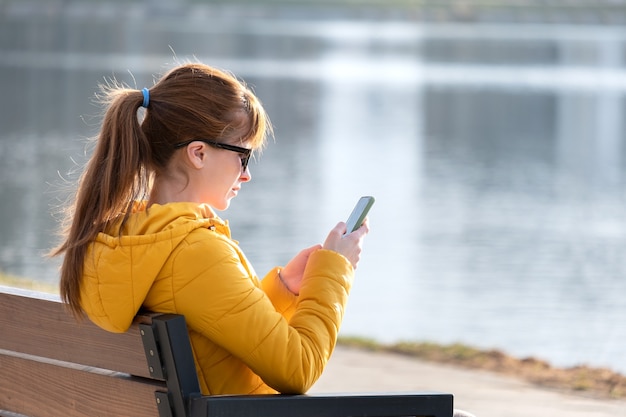 Giovane bella donna seduta su una panchina del parco navigando il suo telefono all'aperto la sera.