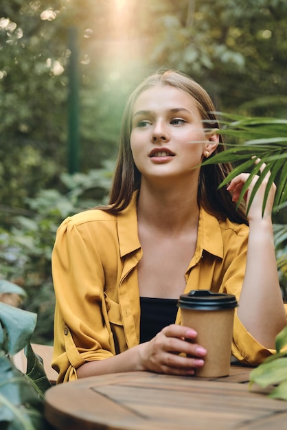 Giovane bella donna sognante in camicia gialla seduta con una tazza di caffè per andare al tavolo di legno con foglie verdi intorno nel parco