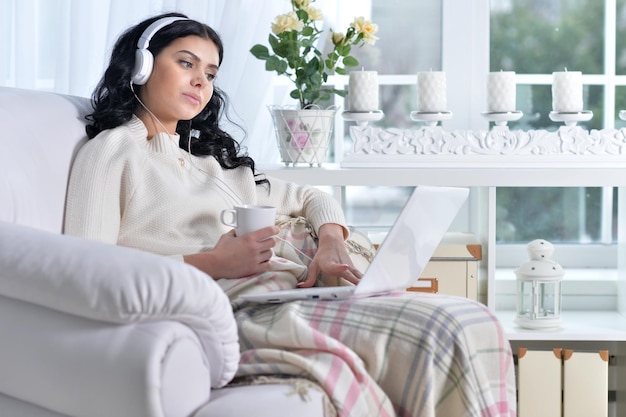 Giovane bella donna sorridente che si siede alla poltrona e che utilizza il computer portatile