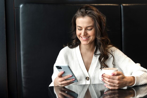 Giovane bella donna sorridente che tiene una tazza di caffè e guarda lo smartphone