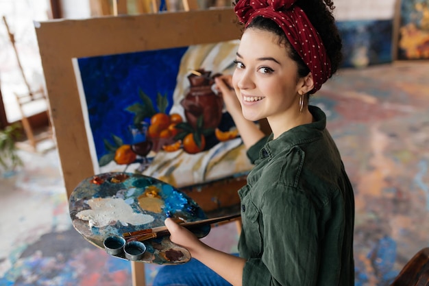 Giovane bella donna sorridente con i capelli ricci scuri seduto su una sedia che disegna un'immagine straordinaria su tela mentre guarda felicemente in camera in un accogliente laboratorio d'arte