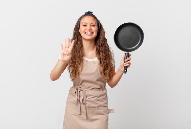 Giovane bella donna sorridente e dall'aspetto amichevole, mostrando il concetto di chef numero quattro e tenendo in mano una padella