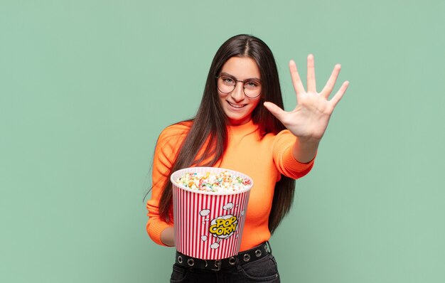 Giovane bella donna sorridente e dall'aspetto amichevole, mostrando il numero cinque o il quinto con la mano in avanti, conto alla rovescia. concetto di pop corn