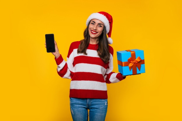 Giovane bella donna sorridente eccitata mentre mostra scatola regalo di Natale e smartphone in mano e posa su sfondo giallo