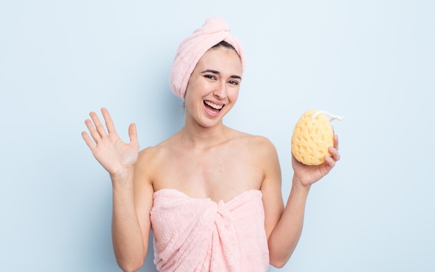 Giovane bella donna sorridente felicemente agitando la mano dandoti il benvenuto e salutandoti doccia e spugna concetto