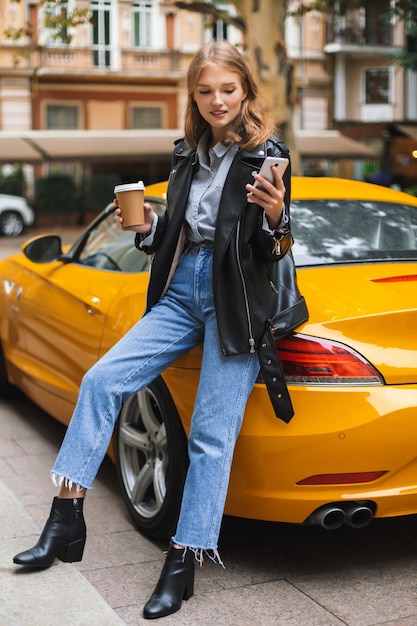 Giovane bella donna sorridente in giacca di pelle che si appoggia su auto sportiva gialla che tiene tazza di caffè per andare in mano mentre sognante utilizza il cellulare sulla strada della città