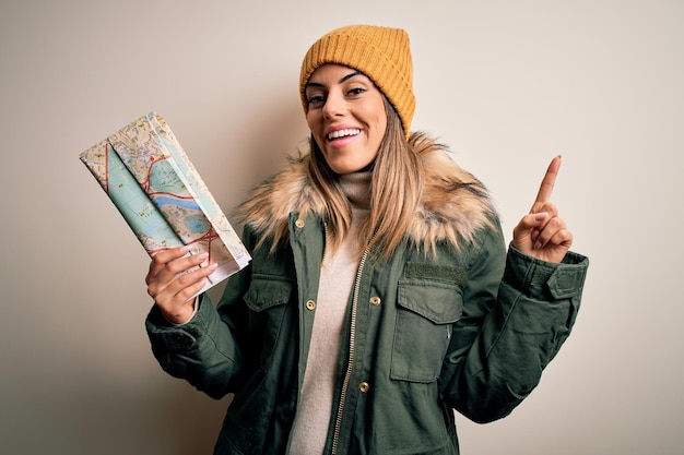 Giovane bella donna turistica che indossa abiti invernali tenendo la mappa della città su sfondo bianco sorpreso da un'idea o una domanda che punta il dito con la faccia felice numero uno