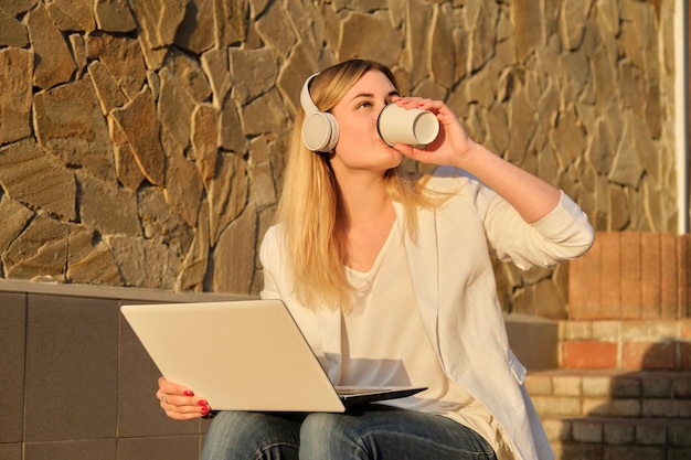 Giovane bella femmina in cuffia con il computer portatile che beve caffè in tazza di carta