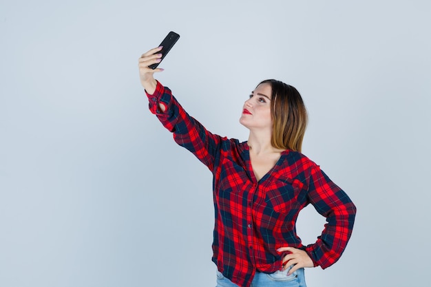 Giovane bella femmina prendendo selfie, con la mano sulla vita in camicia casual e guardando allegro, vista frontale.