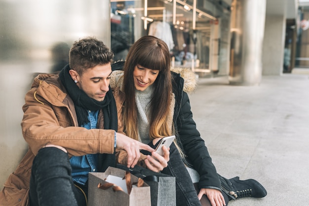 Giovane bella innamorata coppia shopping per la strada prendendo una pausa a sedere sul pavimento lo shopping sul telefono cellulare