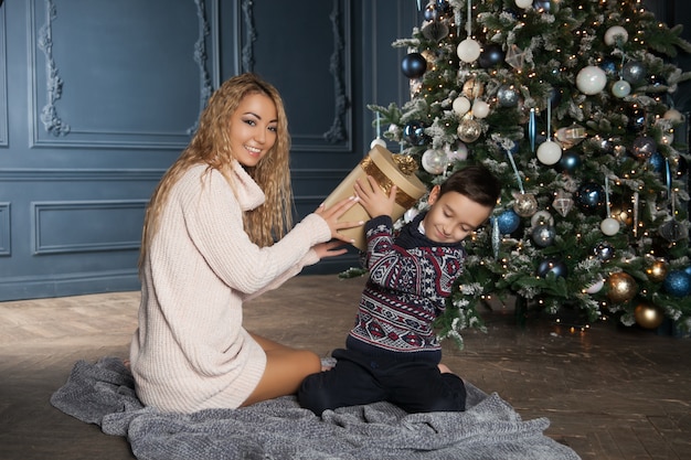 Giovane bella madre asiatica con il suo piccolo figlio seduto vicino a un albero di Natale decorato e regali di scambio.