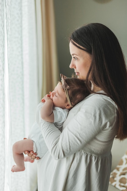 Giovane bella madre dai capelli scuri tiene in braccio la figlia appena nata in una casa accogliente Ritratto di famiglia Maternità Giovane donna bacia il suo bambino di 2 mesi