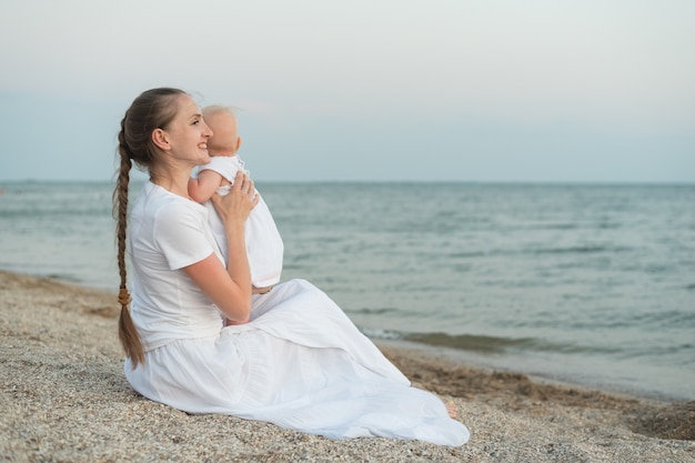 Giovane bella mamma che si siede sulla spiaggia e che abbraccia bambino.