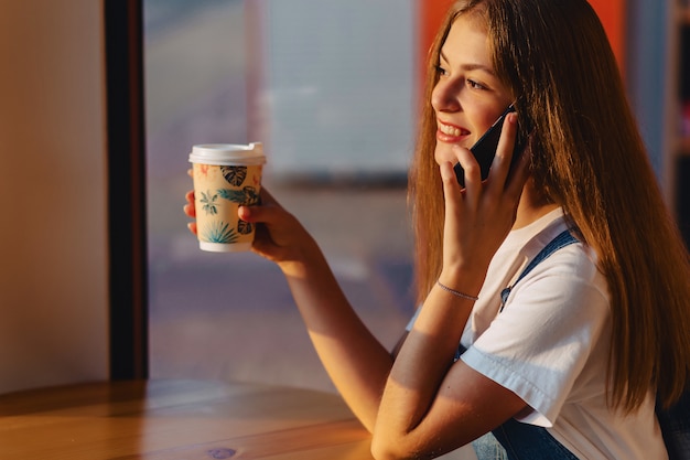 Giovane bella ragazza attraente al caffè con caffè e telefono al mattino travi