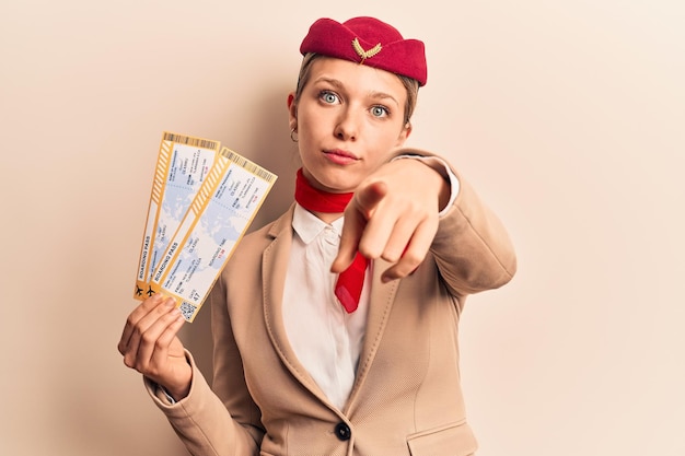 Giovane bella ragazza bionda che indossa l'uniforme da hostess con la carta d'imbarco che punta con il dito verso la fotocamera e ti fa un gesto fiducioso che sembra serio