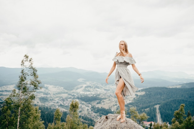 Giovane bella ragazza bionda scalza con capelli lunghi in vestito da estate che sta sopra la montagna alla pietra e che gode della vista scenica del paesaggio favoloso.