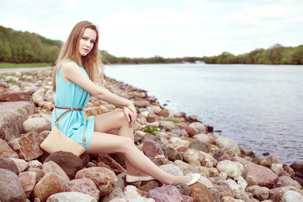 Giovane bella ragazza bionda seduta sulle rocce, sulla riva del fiume.