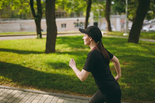 Giovane bella ragazza bruna atletica concentrata in uniforme nera e allenamento con berretto facendo esercizi sportivi in esecuzione guardando dritto sul sentiero nel parco cittadino all'aperto