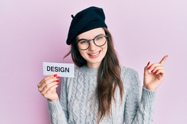 Giovane bella ragazza caucasica che tiene la parola design su carta sorridente felice indicando con la mano e il dito di lato