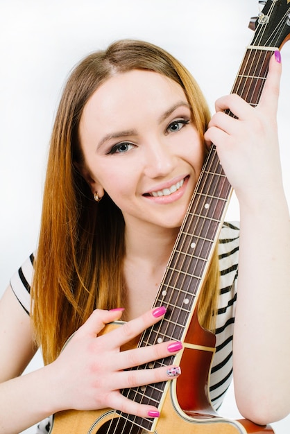 Giovane bella ragazza che indossa jeans e una maglietta a righe con una chitarra acustica su sfondo bianco
