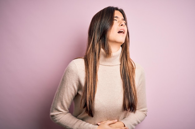 Giovane bella ragazza che indossa un maglione a colletto casuale in piedi su uno sfondo rosa isolato con la mano sullo stomaco perché nausea malattia dolorosa sentimento di malessere concetto di dolore