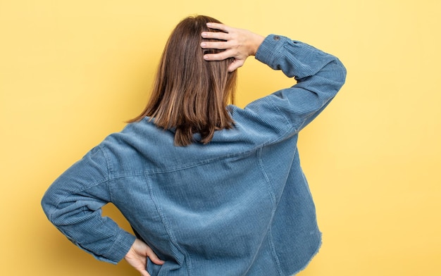 Giovane bella ragazza che si sente incapace e confusa, pensando a una soluzione, con la mano sull'anca e l'altra sulla testa, vista posteriore