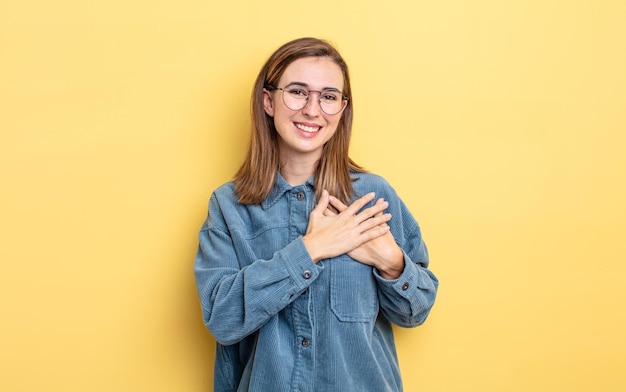 Giovane bella ragazza che si sente romantica, felice e innamorata, sorride allegramente e si tiene per mano vicino al cuore