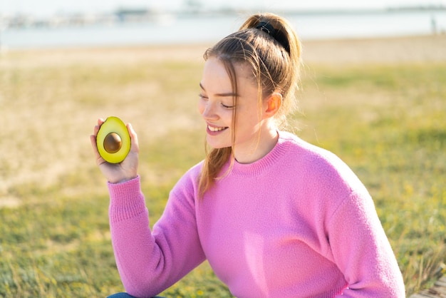 Giovane bella ragazza che tiene un avocado all'aperto con felice espressione