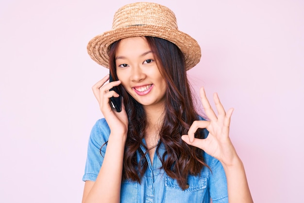 Giovane bella ragazza cinese che indossa cappello estivo che ha una conversazione che parla sullo smartphone facendo segno ok con le dita, sorridente simbolo eccellente gesticolare amichevole