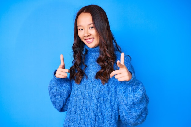 Giovane bella ragazza cinese che indossa un maglione invernale casual che punta le dita verso la macchina fotografica con una faccia felice e divertente di buona energia e vibrazioni