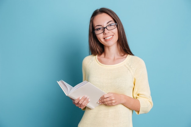 Giovane bella ragazza con gli occhiali che tiene il libro isolato sullo sfondo blu