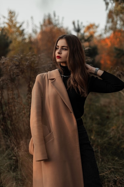 Giovane bella ragazza con le labbra rosse in un cappotto beige alla moda con un maglione nero all'aperto nell'erba